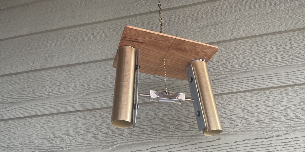 A door chime hanging from a chain against grey siding: two brass tubes under a wooden board, a striker on a bar between them
