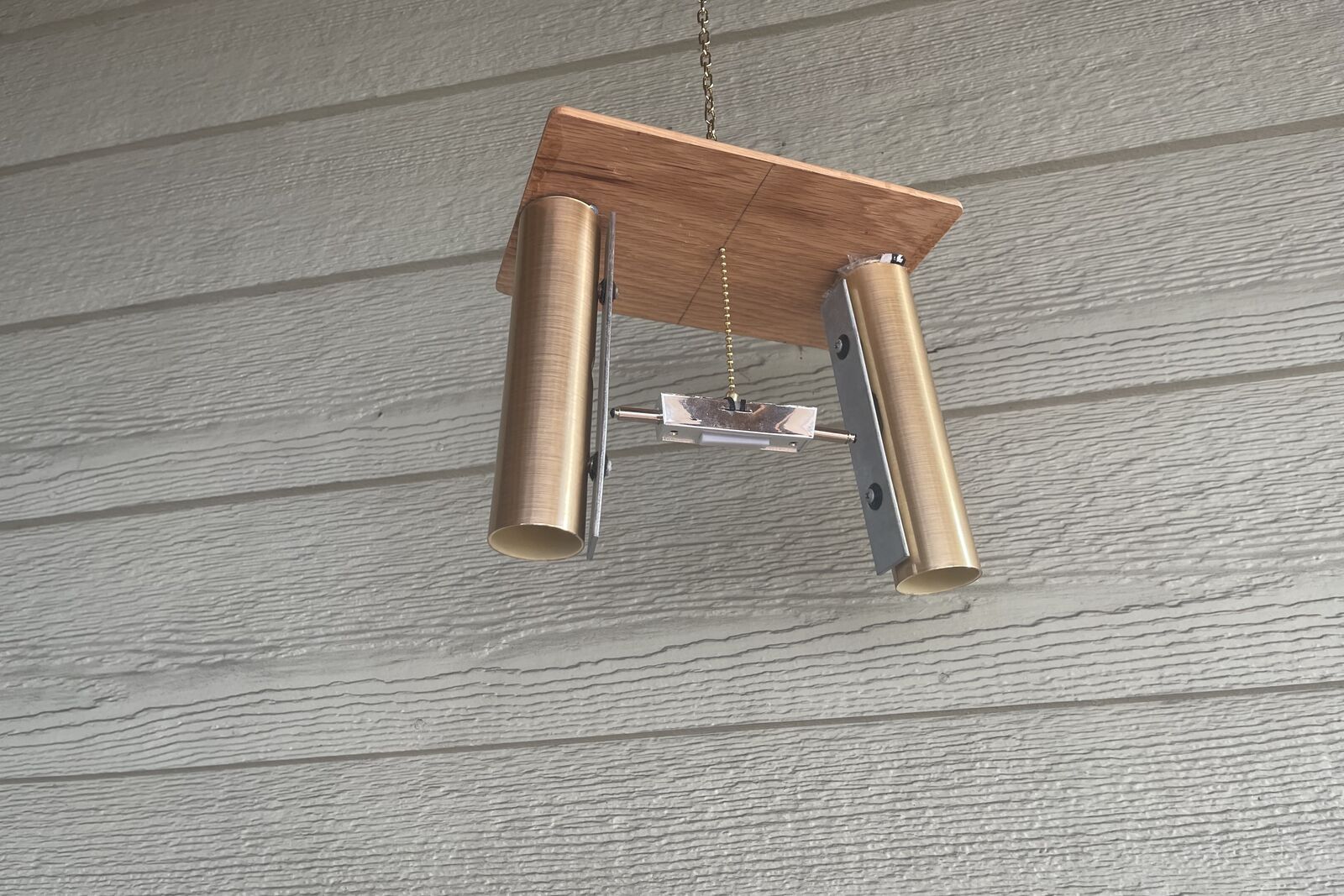 A door chime hanging from a chain against grey siding: two brass tubes under a wooden board, a striker on a bar between them