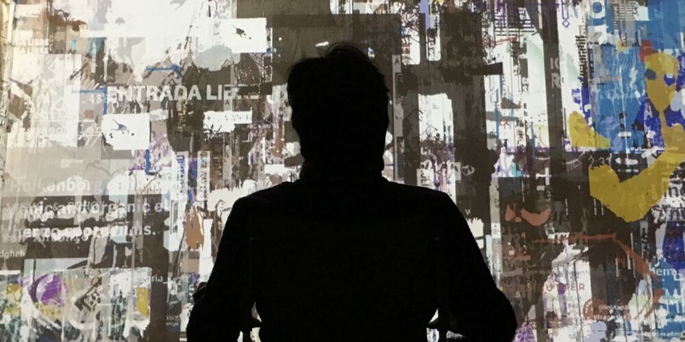 A person in silhouette, seated in front of a wall-sized projection of torn and layered images and text