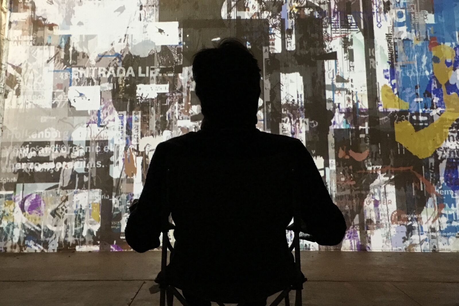 A person in silhouette, seated in front of a wall-sized projection of torn and layered images and text