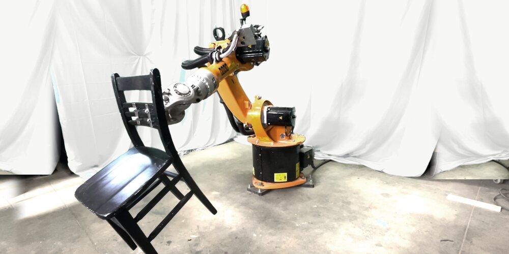An orange KUKA robotic arm gripping the back of a black wooden chair and tilting it, in front of a white curtain (edited: the green screen behind it was recolored white)