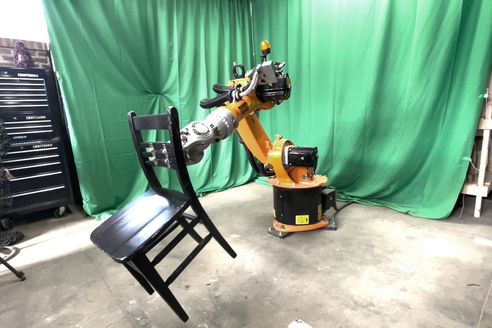 An orange KUKA robotic arm gripping the back of a black wooden chair and tilting it, in front of a green screen