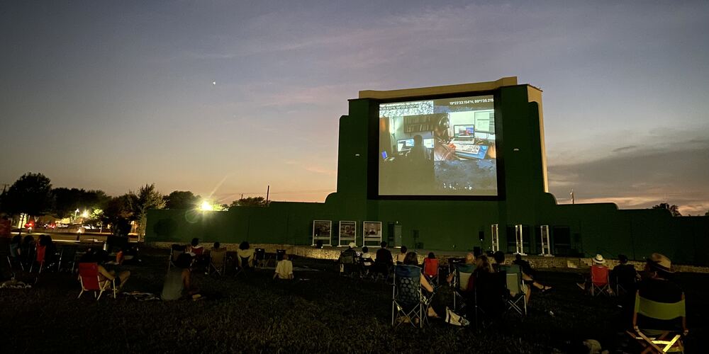 The outdoor screening at Mission Marquee Plaza at dusk: the old drive-in screen shows the video, an audience in lawn chairs on the grass in front
