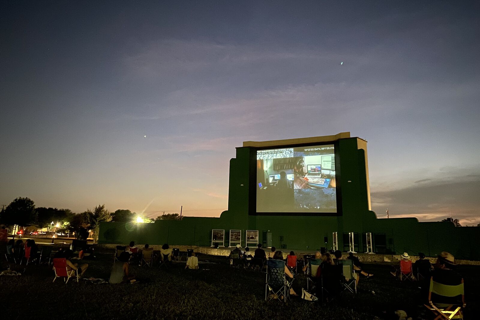 The outdoor screening at Mission Marquee Plaza at dusk: the old drive-in screen shows the video, an audience in lawn chairs on the grass in front