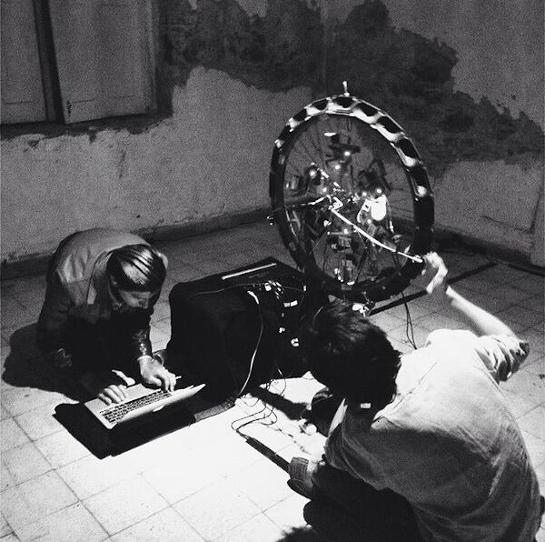 In black and white, two people on the floor working at a laptop beside the wheel