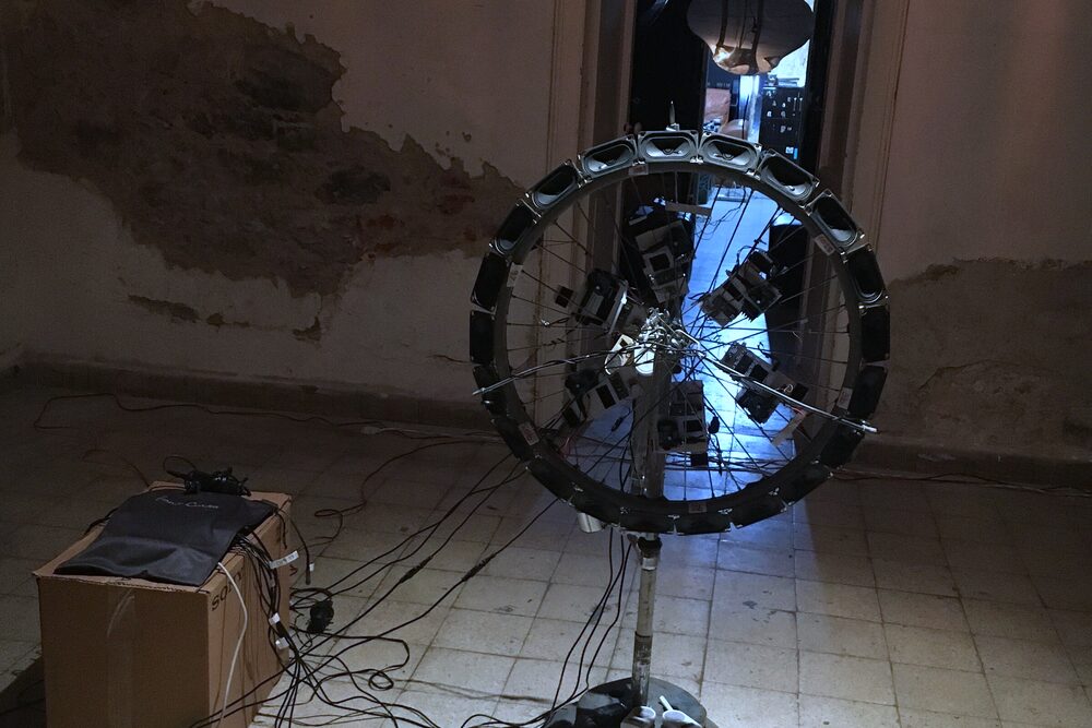 A bicycle wheel lined with small speakers, standing on a pole in a room with peeling walls, cables running to a box on the floor