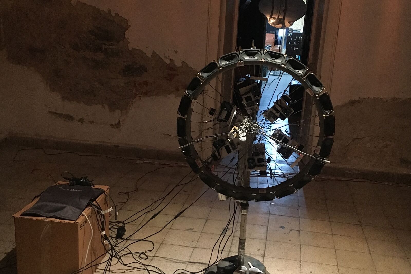 A bicycle wheel lined with small speakers, standing on a pole in a room with peeling walls, cables running to a box on the floor
