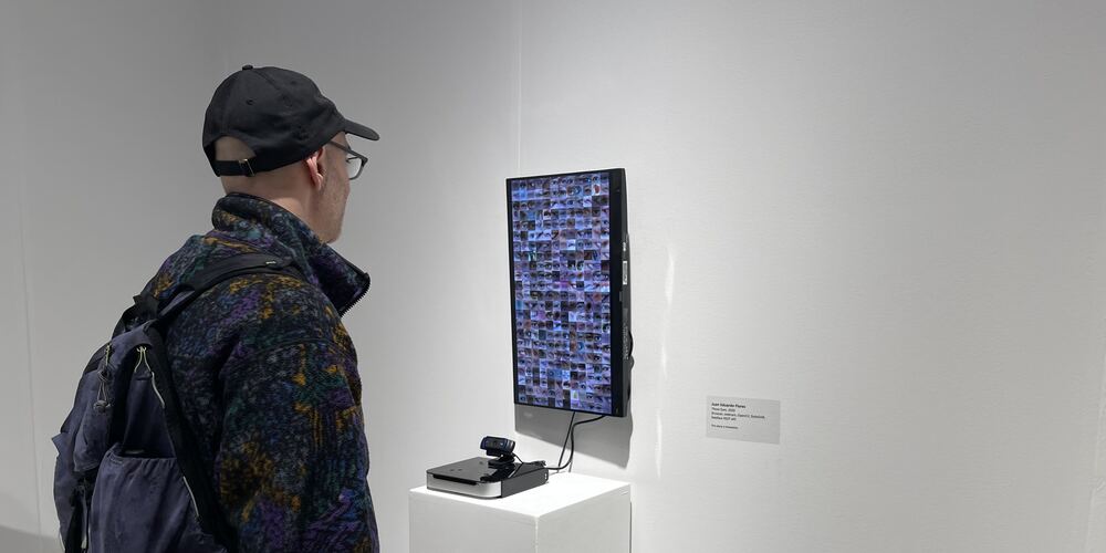 A visitor looking at a vertical monitor filled with a grid of cropped eyes, a webcam on the plinth below