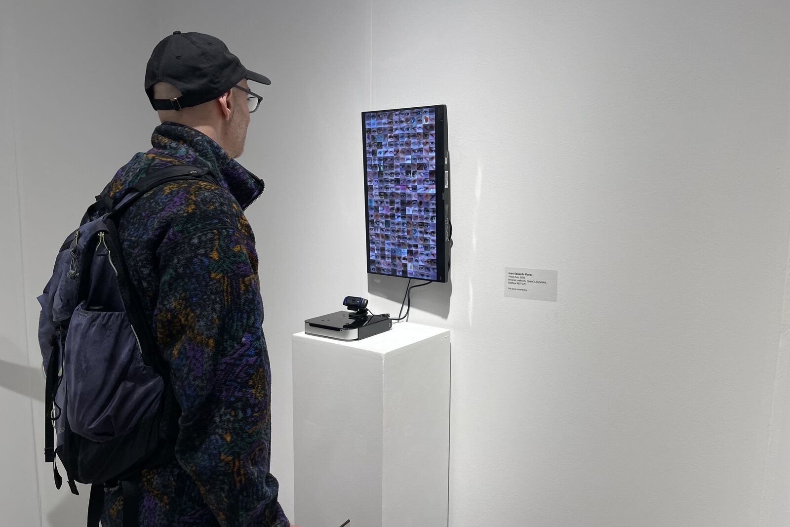 A visitor in a cap and backpack looking at a vertical monitor filled with a grid of cropped eyes, a webcam on the plinth below
