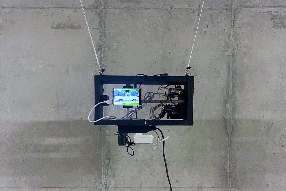 A black frame hung by two cords on a concrete wall, holding a small screen that shows a white pickup truck