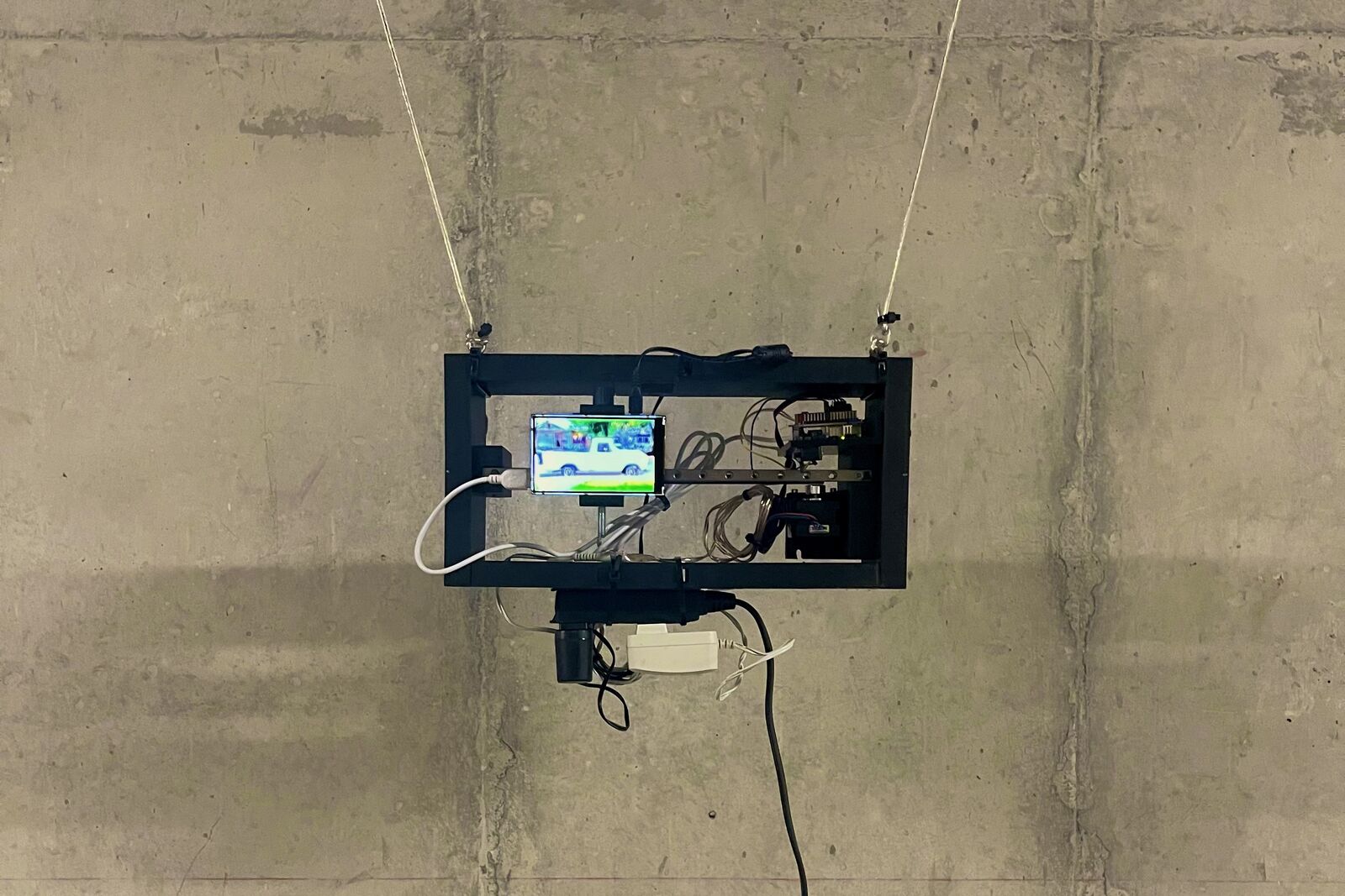 A black frame hung by two cords on a concrete wall, holding a small screen that shows a white pickup truck, a metal rail and exposed wiring